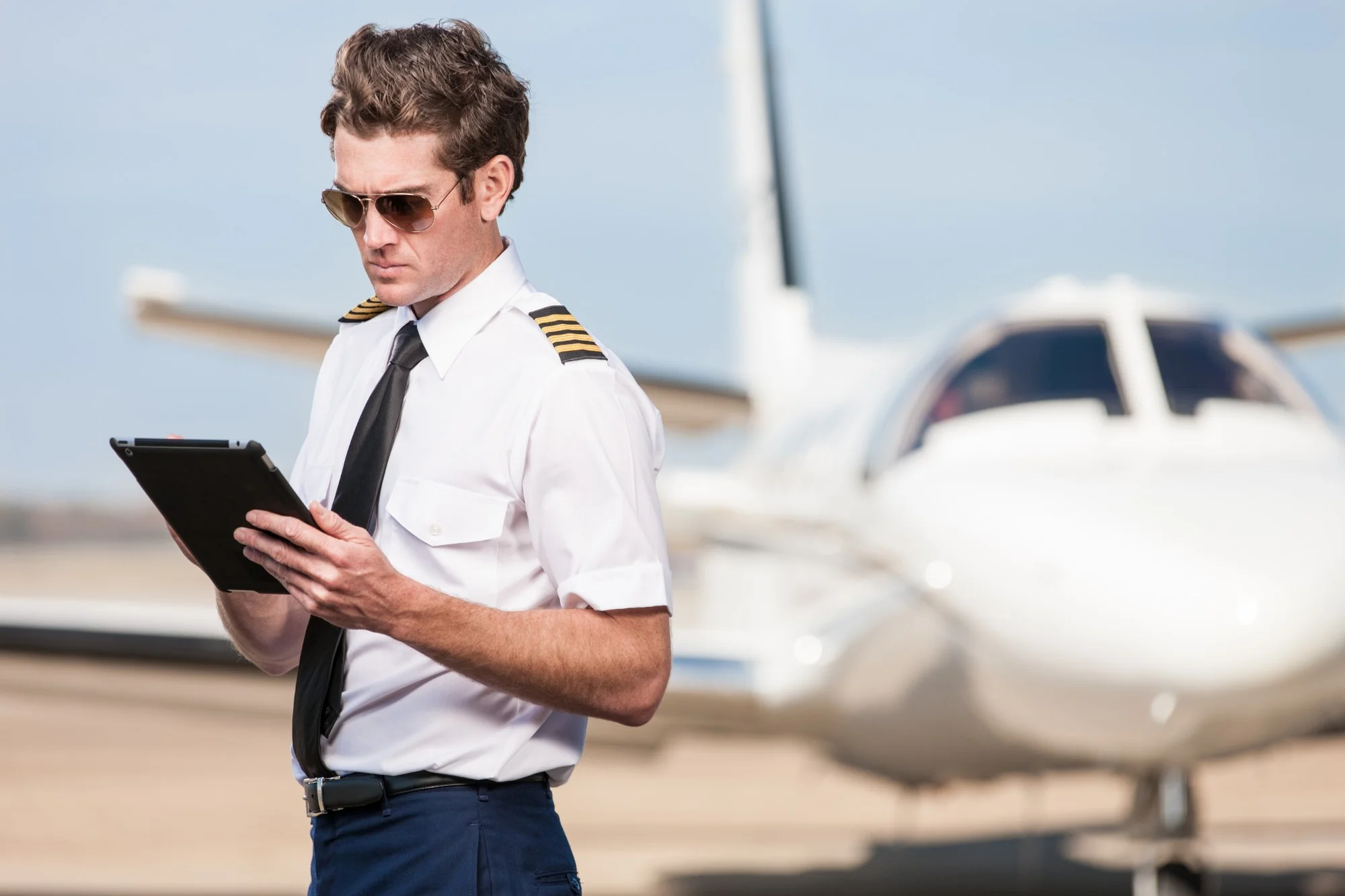 A captain in sunglasses reading a tablet on the ramp, a business jet parked behind him